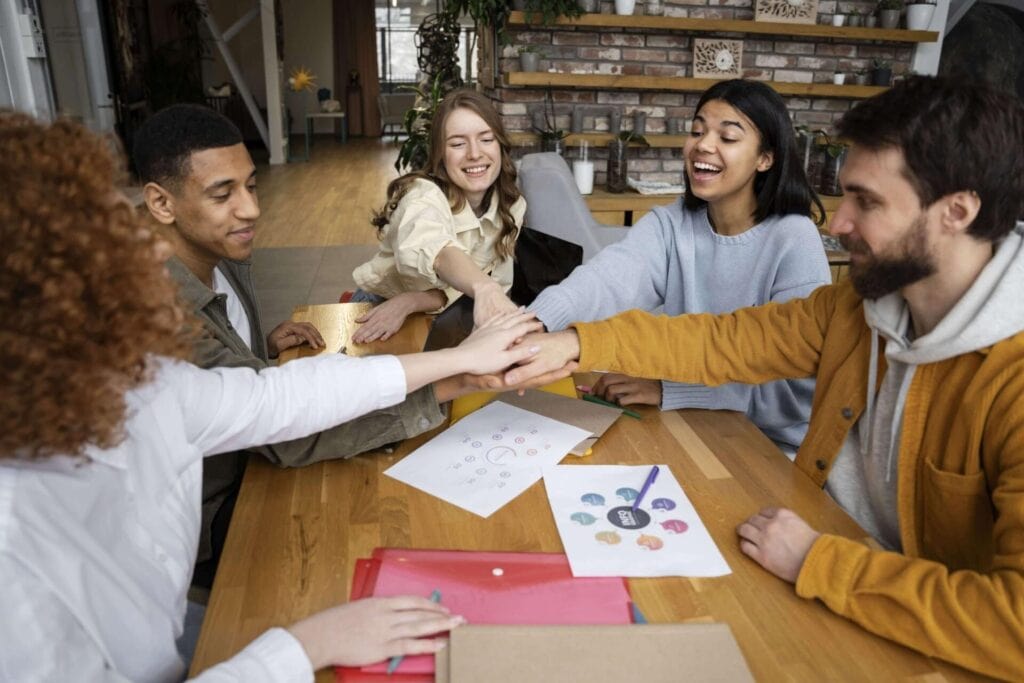 Collaborative team smiling and placing hands together at a wooden table in a modern office setting, happy that they now have an employee benefit plan for their team.